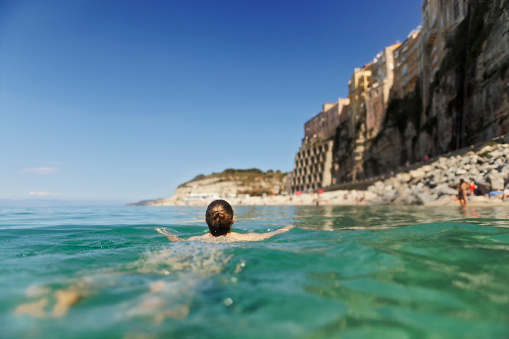 Teenage girl enjoying sunny spring day in Calabrian town of Tropea. She is swimming in cold water. Behind her there are stunning cliffs with buildings of the small Italian town Tropea.\nSpringtime, off-season vacations day in Tropea, Catania, Italy.\nShot with Canon R5