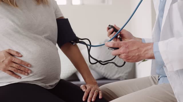 Caucasian doctor testing blood pressure of pregnant Caucasian female. Daytime At home doctor consultation in modern home