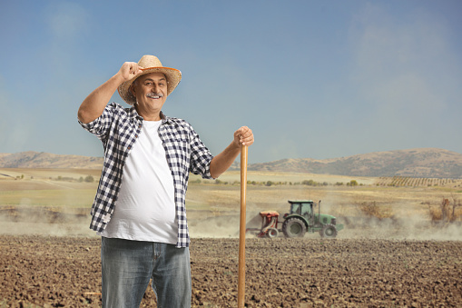 Portrait of a happy mature farmer with a shovel standing on a dusty field with tractor