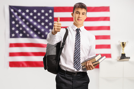 Teenage male student holding books and showing thumbs up in front of a USA flag