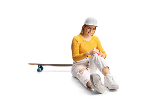 Young female with a skateboard sitting on the floor and holding her injured knee isolated on white background