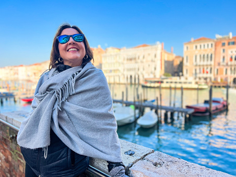 Winter in Venice, Italy concept: Travel and vacation destination background. Female tourist with mirror sunglasses posing in a pashmina in front of the Grand canal in Venice on a cold sunny day