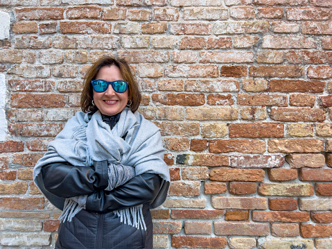 Winter in Venice, Italy concept: Travel and vacation destination background. Female tourist with mirror sunglasses posing in a pashmina in front of a brown brick wall.