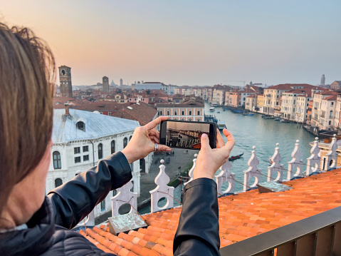 Winter in Venice, Italy concept: Travel and vacation destination background. Woman taking photo of the beautiful Grand Canal over the Rialto Bridge at the roof terrase of T Fondaco Dei Tedeschi mall at Sunset, a breathtaking view, picturesque scene.