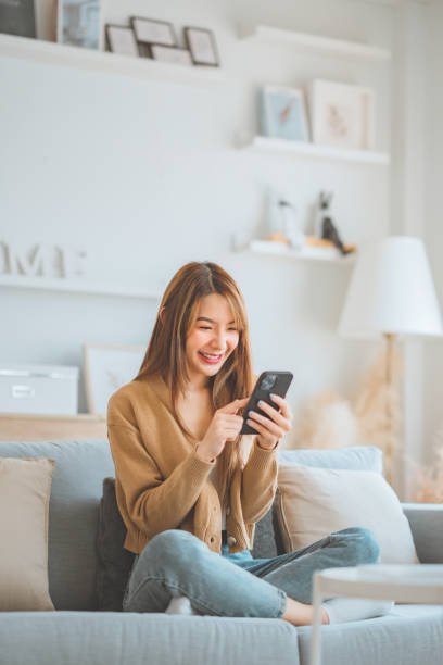 istock Happy young asian woman browsing surfing wireless internet on mobile phone while sitting a couch in living room at home. 1489956049