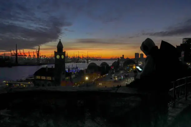 Silhouette of a teenager sitting on a wall looking at his glowing smartphone, in the background the port of Hamburg at sunset, horizontal