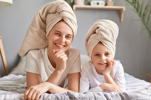 Beauty portrait of happy mom and daughter in towels on head lying on bed at home, posing after bath, having spa day, cute little girl and her mother having fun together.