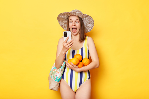 Surprised excited woman wearing striped swimsuit and straw hat isolated yellow background standing with fruits using mobile phone for booking tickets.
