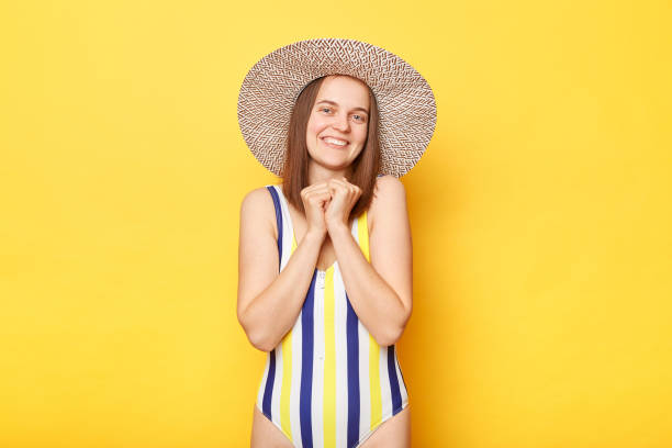 istock Woman with cheerful expression wearing striped swimsuit and sun hat while standing isolated on yellow background, looking directly at the camera, enjoying peaceful day at beach. 1489968559