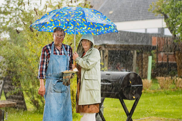 Rainy Day BBQ: Disappointed Senior Couple Under Umbrella A senior man and woman make disappointed faces as they stand under an umbrella in front of their still-smoking barbecue during a countryside event, caught in unexpected rain. He wears a checkered shirt and apron, while she dons a raincoat. unusual couple stock pictures, royalty-free photos & images