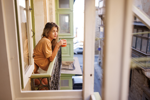 Young beautiful woman standing on balcony and drinking coffee in the morning
