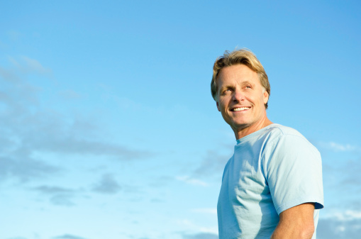 color portrait photo of a happy smiling blond haired man in his forties wearing a blue t'shirt against a blue sky backround.
