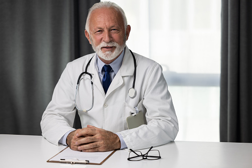 Portrait of senior male doctor, sitting by the desk and looking at a camera. Medical director professional of hospital wearing uniform and showing confidence