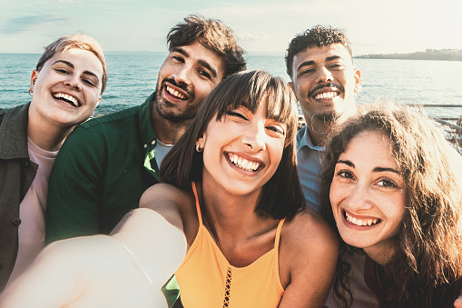 A diverse group of friends, including a non-binary person, take a selfie on the waterfront on a warm, sunny day. The five young adults, all in their twenties, smile at the camera