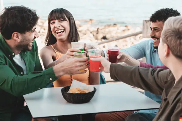 Group of five friends toasting with colorful fizzy drinks on a seaside terrace, enjoying a summer gathering - summertime people lifestyle