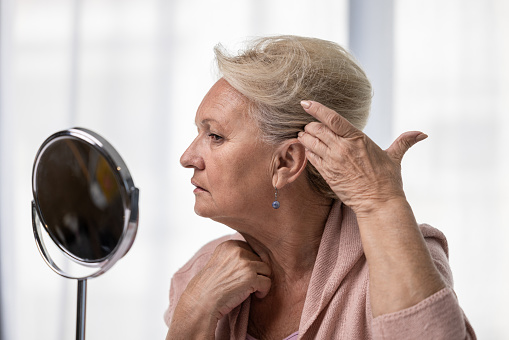 Elderly woman checking her hairline or grey hairs by looking at mirror. Alopecia, hair loss and aging concept.