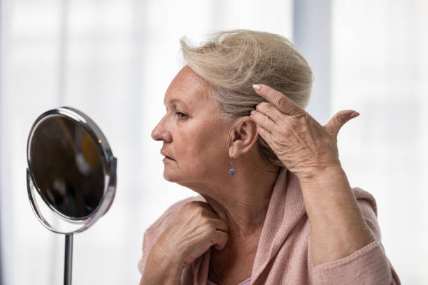 Senior woman checking her hairline or grey hairs by looking at mirror. Alopecia, hair loss and aging concept. Elderly woman checking her hairline or grey hairs by looking at mirror. Alopecia, hair loss and aging concept. woman hairline stock pictures, royalty-free photos & images