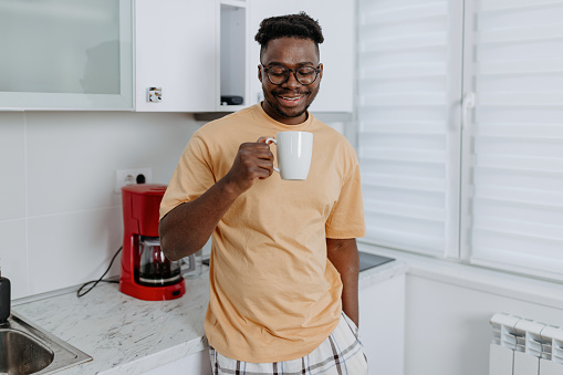 A young African American man in pajamas stands in his kitchen, holding a cup of coffee