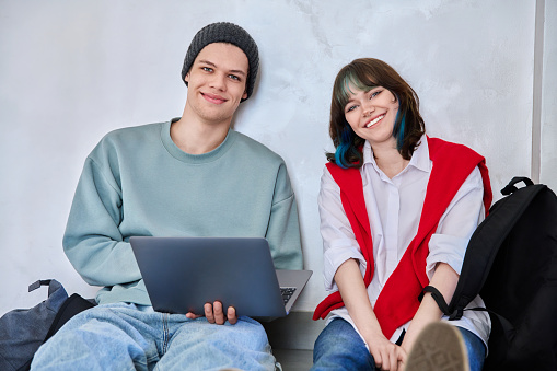 Friends hipster teenagers guy and girl sitting on floor with backpacks using laptop looking at camera inside study hall corridor. Fashion trendy youth, lifestyle, communication, friendship concept