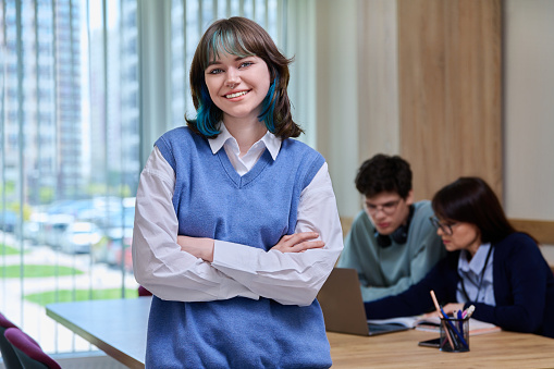 Portrait of smiling trendy female student 18, 19 years old looking at camera, posing in college classroom, education center. Confident teenage female with crossed arms. Education, knowledge, learning