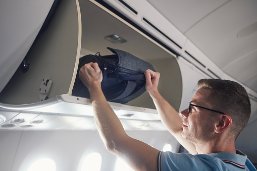 Man travel by airplane. Passenger putting hand baggage in lockers above seats of plane.