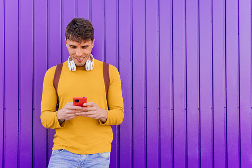 Young hispanic man in yellow clothes on purple background, using his smart phone outdoors. Person who uses social networks with his mobile phone.
