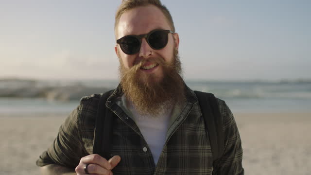 bearded hipster man with tattoos posing confidently at beach wearing sunglasses smiling happy
