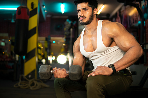Shot of serious, muscular Indian young man doing biceps curls exercise with dumbbells at the gym.