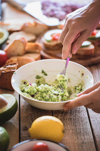 Guacamole preparing, raw avocado with chopped red onions smashed in bowl using fork, close-up