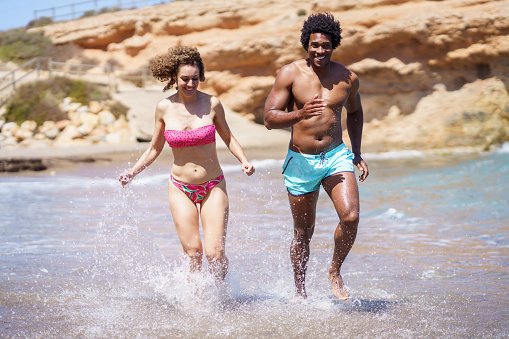 Full body of young African American male looking at camera, while running with looking down diverse female in splashing water of waving sea during sunny day