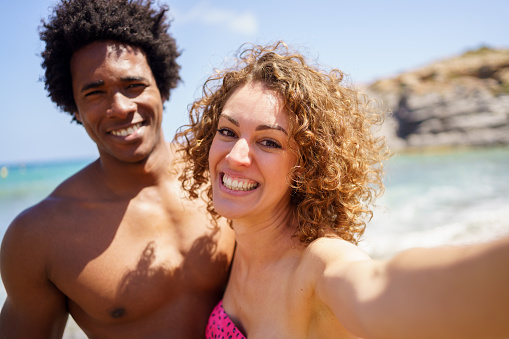 Cheerful multiracial girlfriend and boyfriend in swimwear smiling while taking selfie on sunny day near sea during summer holidays