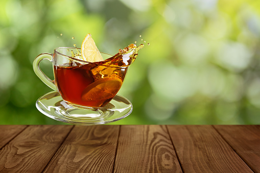 flying glass cup of tea with splash and falling slice of lemon on wooden table with green blurred background