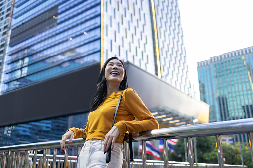 Young Asian woman in yellow shirt standing on footbridge looking at cityscapes in front of Krungsri bank office