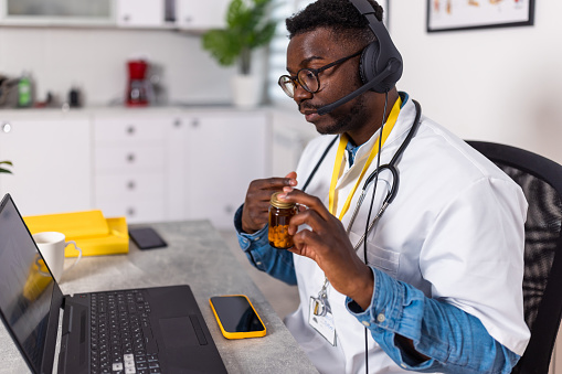 A young African American male doctor is sitting at his office desk and holding a bottle of pills, while talking to his patient in a video call.