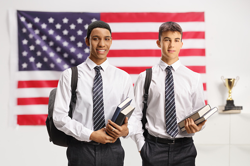 Caucasian and african american male students in a shirt and tie holding books and standing in front of a USA flag