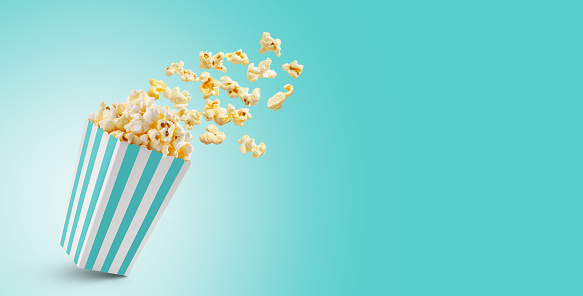 Popcorn flying out of turquoise white striped paper box isolated on turquoise background with copy space. Splash, levitation of popcorn grains.