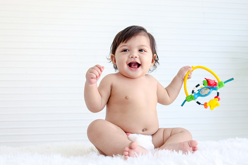 Portrait of smiling crawling baby sits on fluffy white rug, little cute kid girl playing with developmental toys, happy daughter child playing and laughing, love in family, childhood care and learning
