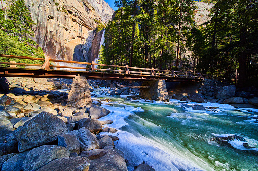 Image of Bridge crossing over dangerous slushy cold river with Yosemite Lower Falls in background and cliffs