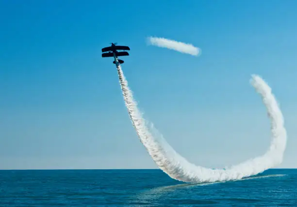 Aerobatic biplane flying with smoke over the sea.