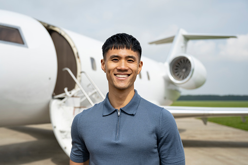 Early 30s man in casual shirt standing on airport tarmac in front of private aircraft and smiling at camera.