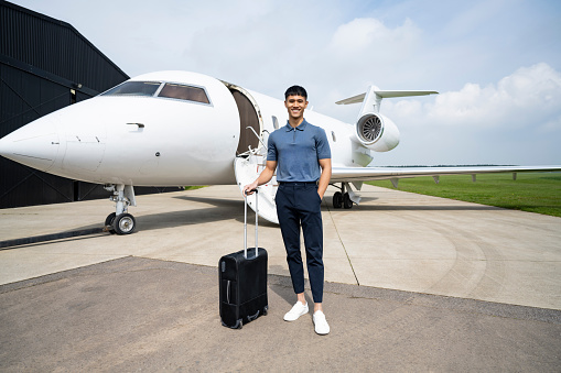 Full length front view of early 30s man in casual attire standing with wheeled luggage, hand in pocket, and smiling at camera with private jet in background.