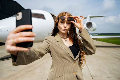 Waist-up view of well-dressed professional in early 20s standing on airport tarmac, lifting her sunglasses, and having fun capturing a memory with smart phone.
