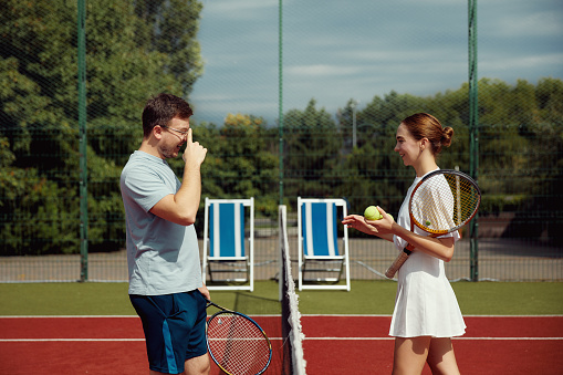 Young tennis players standing and smiling before tennis match. Cheerful man and girl holding rackets in court