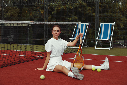 Young beautiful sportswoman with tennis racket sitting at tennis net on tennis court. Sports Fashion. Weekend and sunday activity for recreation