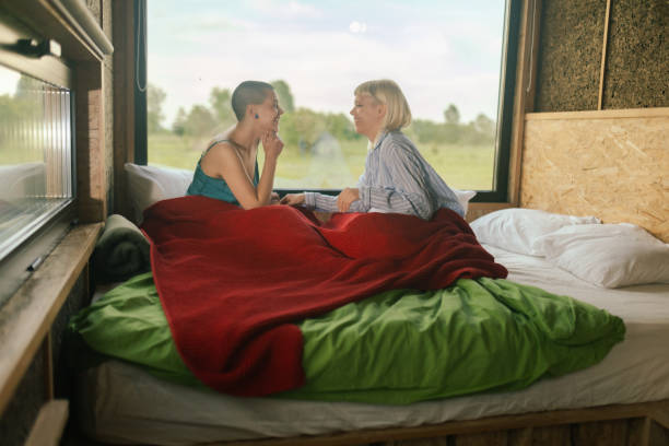 Two lesbian women dressed in pajamas sit in bed by the window in a glamping cabin and enjoy their mornings together. Iceland stock photo