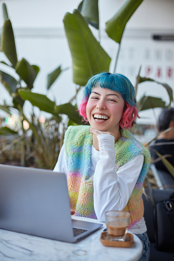 Smiling young woman with colorful hair working with laptop computer in cafe.
