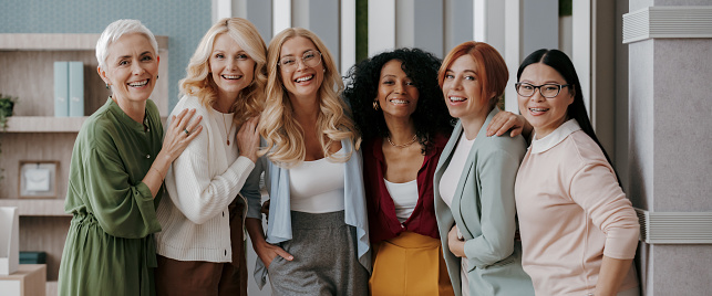 Multi-ethnic group of happy mature women bonding while standing in office together