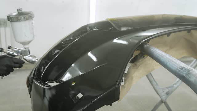Applying paint to the bumper. Paint the bumper with varnish using an airbrush. Close-up of a spray gun being used by a mechanic while painting a car bumper in a spray booth.