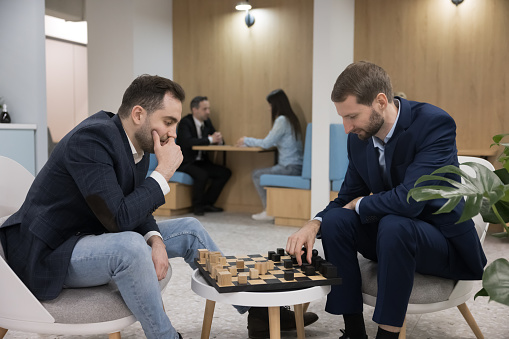 Two male business colleagues playing chess on work break, sitting at chessboard in office kitchen, cafe, enjoying fighting, competition, training intelligence, thinking, moving pieces