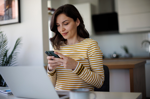 Young smiling businesswoman using a cellphone while working on a laptop in home office